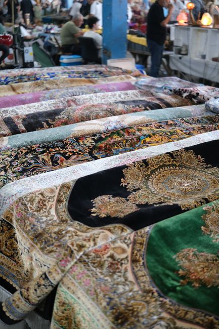 A display of intricately patterned, woven carpets laid out on tables at Brixton Market, with vibrant colors and detailed floral and medallion designs. The carpets' surfaces appear clean and well-maint
