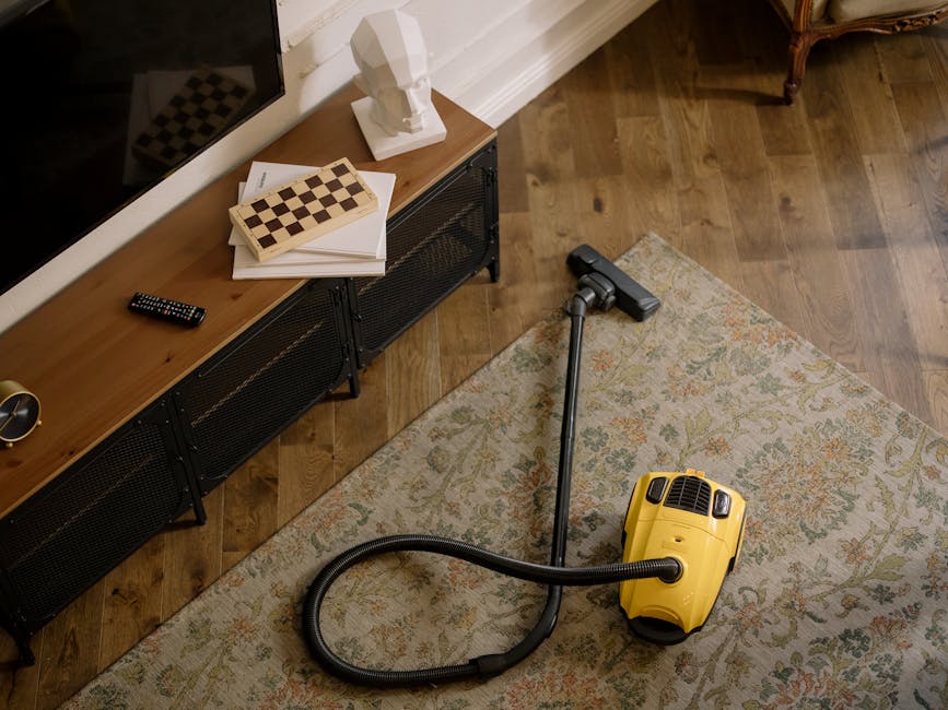Close-up image of a handheld vacuum cleaner with a black body and a slim nozzle attachment, being used on a patterned carpet with floral and geometric motifs in shades of beige, green, and orange. The